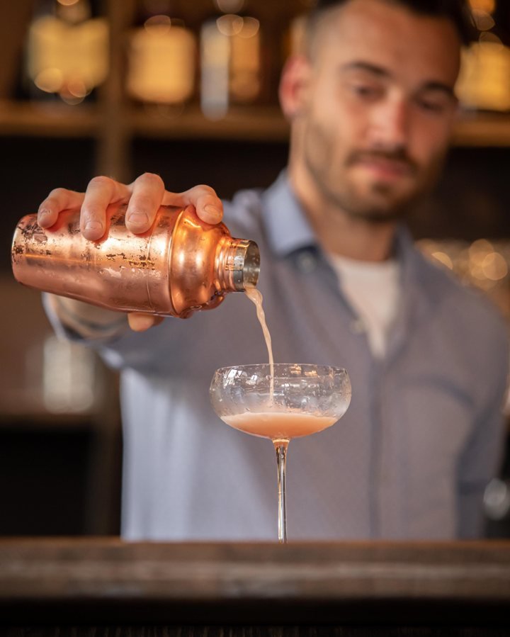 Elvin Angelaud derrière le bar, versant un cocktail au shaker cuivré